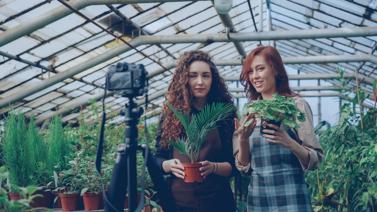 gallery-4 Two women filming a gardening video in a lush greenhouse surrounded by plants.
