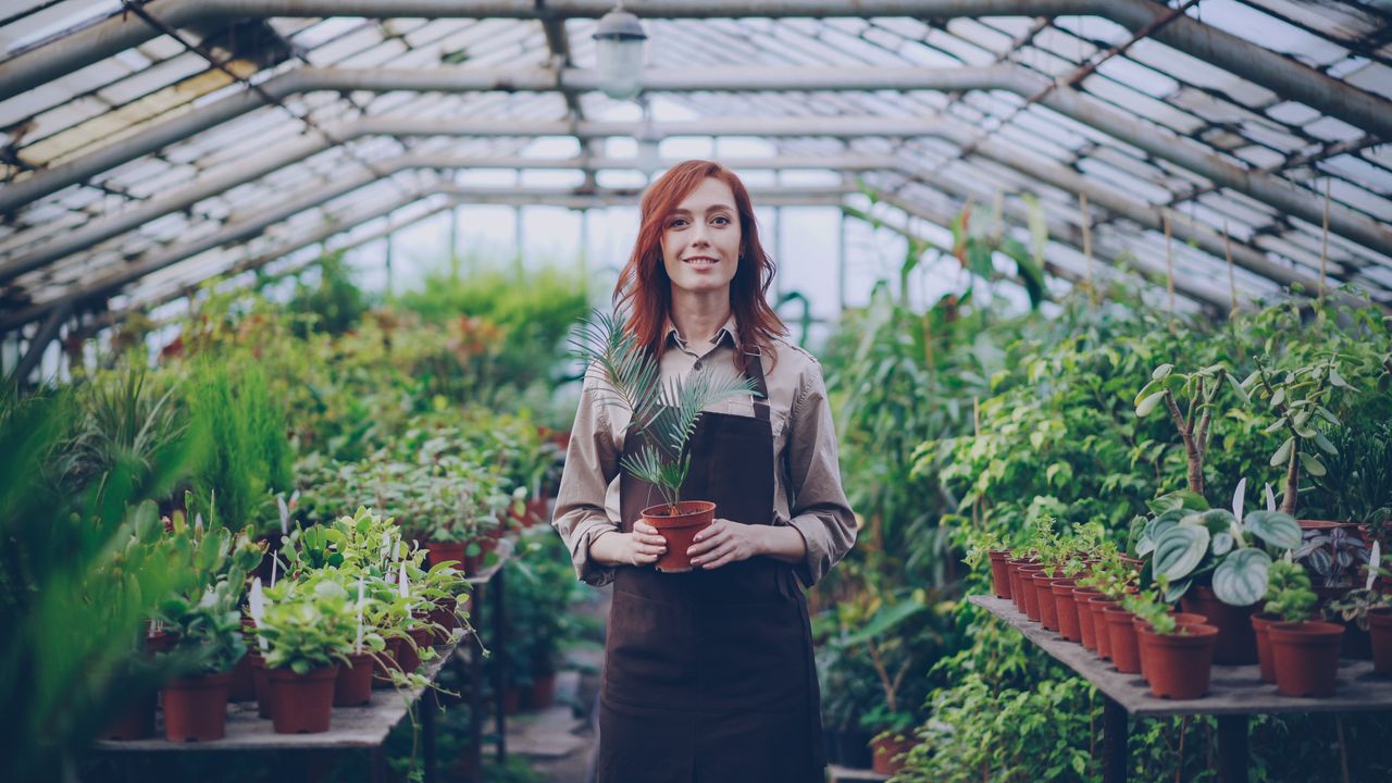 hero-img-02 Woman standing in a lush greenhouse filled with various plants, holding a potted plant.